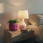Pink Firkle planter with a leafy plant on a bedside nightstand beside a warm lamp and a book in a cozy bedroom