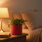Red Orin planter with a trailing pothos plant on a bedside nightstand beside a warm lamp and a book in a cozy bedroom
