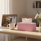A pink ribbed tissue box holder filled with white tissue paper sits on a light wood desk in a bright home office setting, alongside an open laptop, a white mug, and a small vase of daisies.